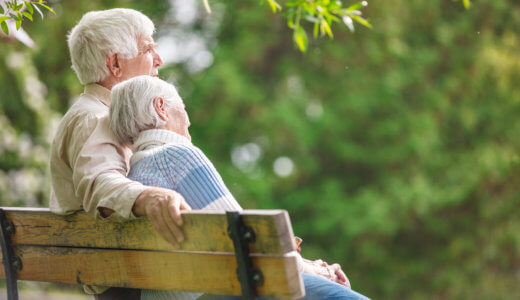 Senior woman rests head on senior man's shoulder as they sit on a park bench