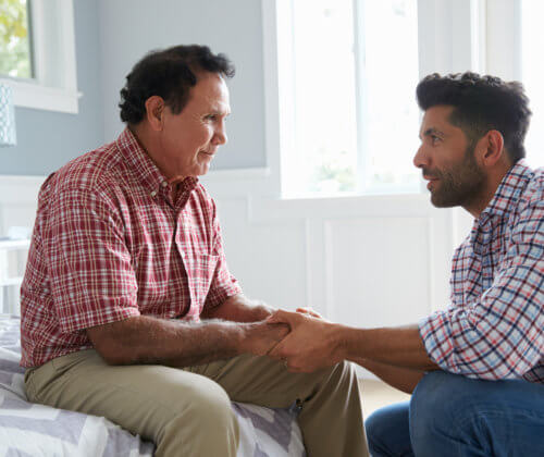 Man sitting on a bed, holding hands with younger man who is kneeling